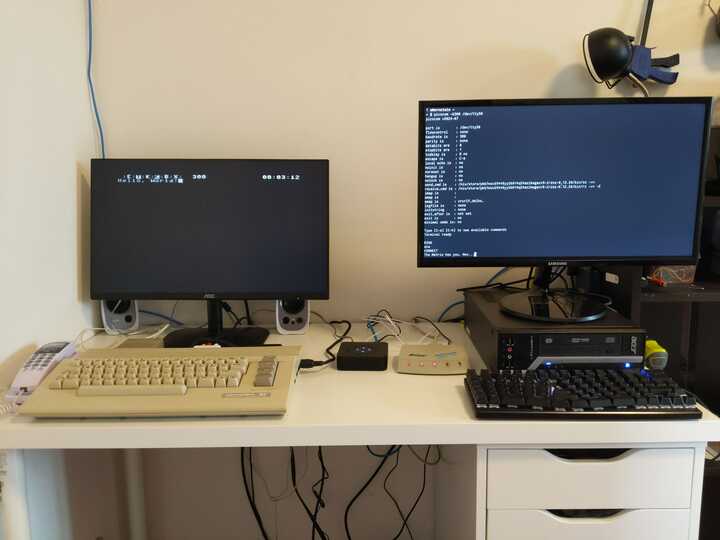 Desk with a pair of computers. On the left is a Commodore 64 and on the right is a PC. Between them is an analog telephone adapter and dial-up modem